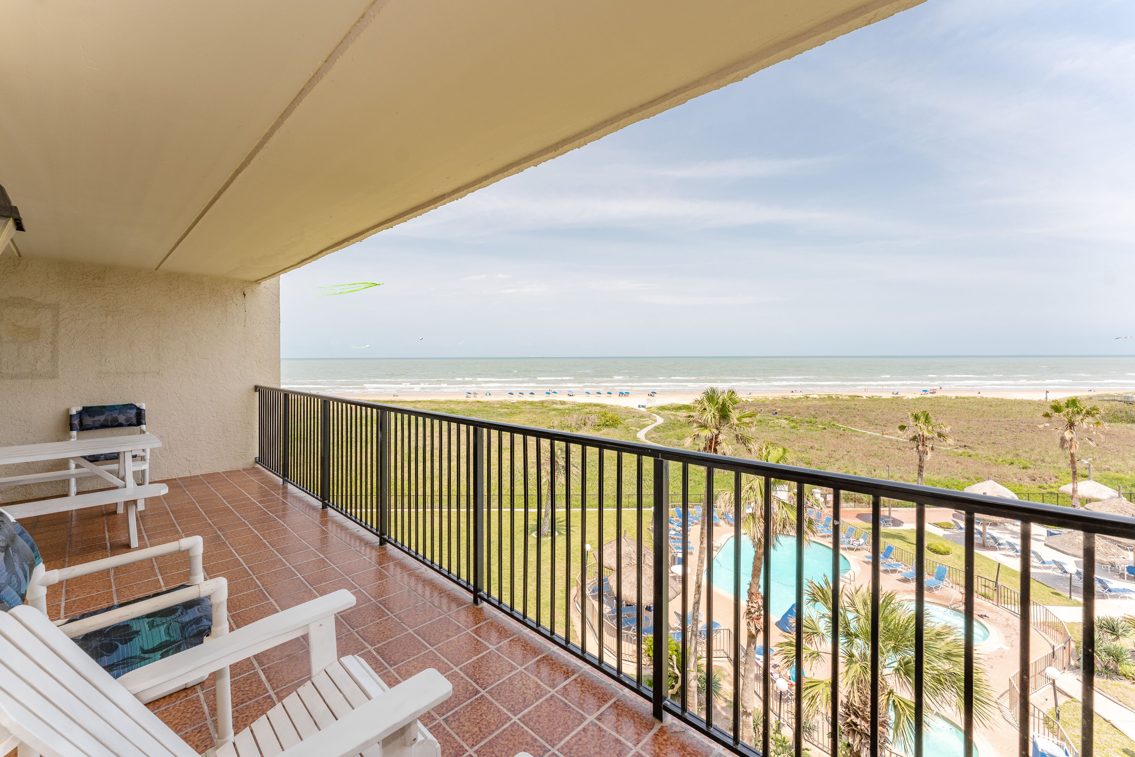 A balcony with white chairs and a table, overlooking a beach and a pool area.