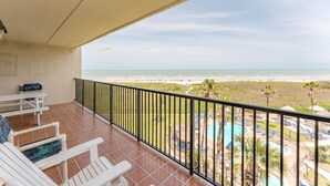 A balcony with white chairs and a table, overlooking a beach and a pool area.