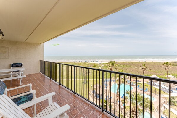 A balcony with white chairs and a table, overlooking a beach and a pool area.