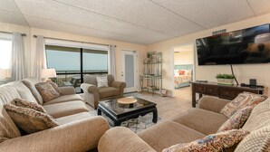 A spacious living room with a large flat-screen TV, comfortable seating, and a balcony view.
