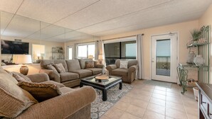 A living room with two sofas, a coffee table, and a TV. There are windows and a door leading to a balcony.