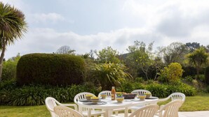 A round outdoor table with white plastic chairs, surrounded by lush greenery and a well-maintained garden. - Les Agapanthes by Interhome (Dinard)