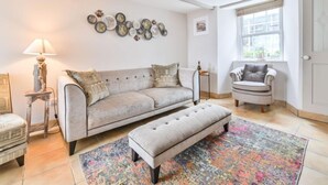 A living room with a grey sofa, a matching ottoman, a patterned rug, a side table with a lamp, and a decorative wall clock. - Blue Crab by Interhome (Mevagissey)