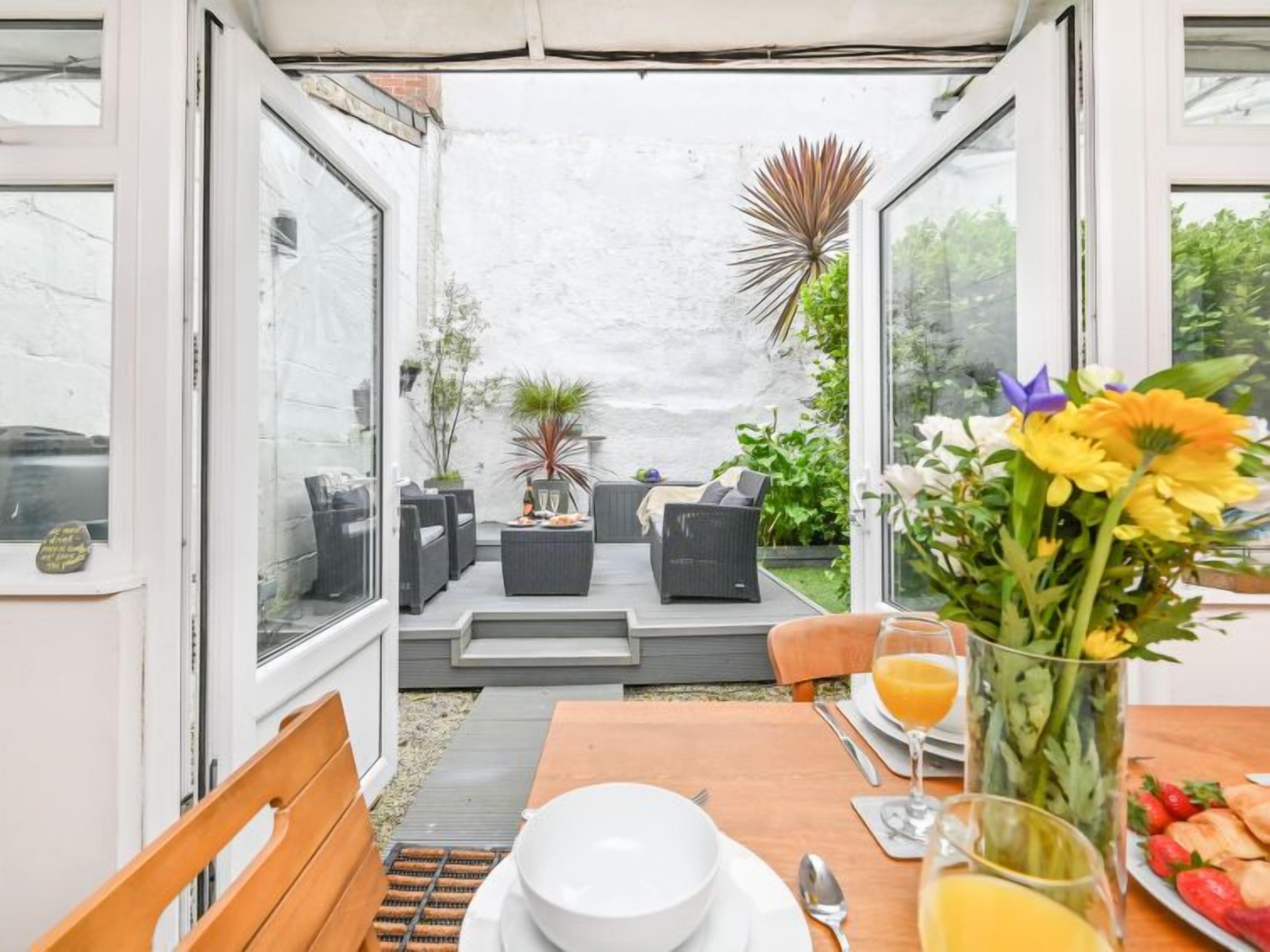 A sunlit patio with outdoor seating, a dining table set for two, and a variety of plants and flowers.