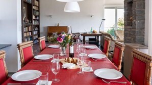 Une salle à manger avec une table rouge dressée pour six personnes, des chaises blanches, un lustre, une bibliothèque et une cheminée en pierre.