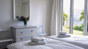 Une chambre d’hôtel avec deux lits, une commode, un miroir et une fenêtre donnant sur un paysage verdoyant et des montagnes.