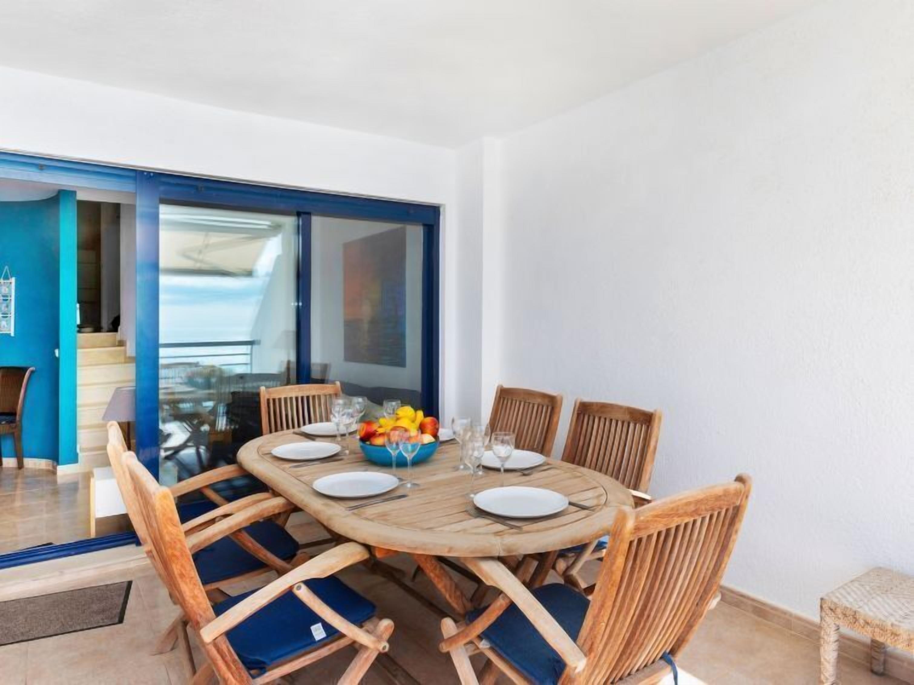 A round wooden dining table with six chairs, a bowl of fruit, and wine glasses set for six.