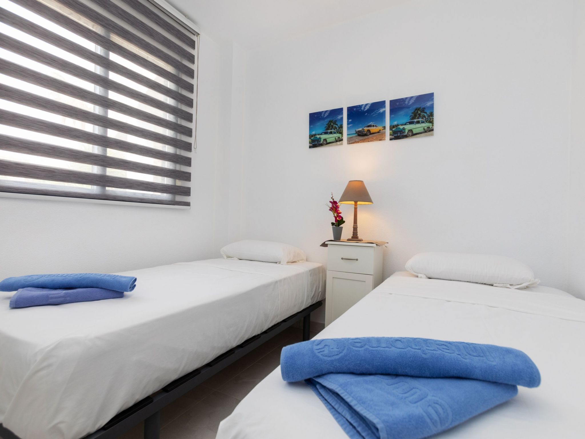 Hotellihuone, jossa on kaksi sänkyä, yöpöytä lampun kanssa ja ikkuna, jossa on sälekaihtimet.
