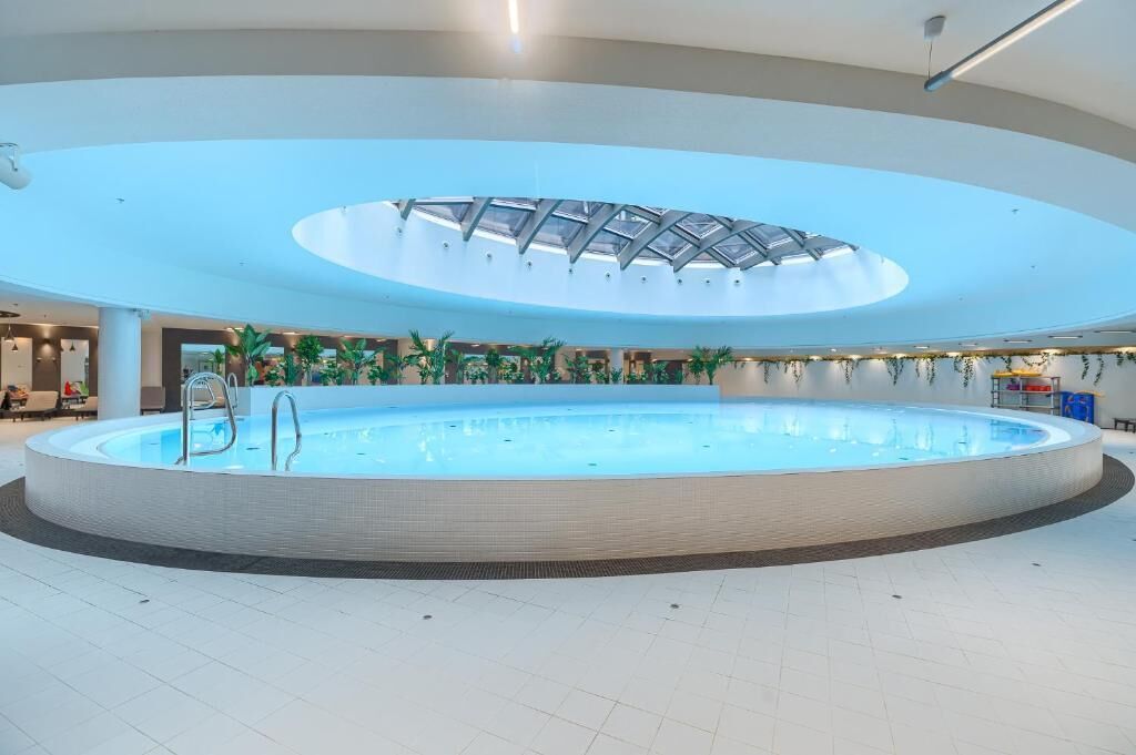 Ein großer Innenpool mit Glasdecke, umgeben von gefliestem Boden in Weiß und Zimmerpflanzen.
