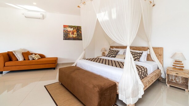 A bedroom with a four-poster bed, a sofa, a nightstand, and a wall-mounted air conditioner. - Luxurious 4BR Villa With Pool, Bali Villa 2206 (Seminyak)