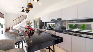 A modern kitchen with a black countertop, white cabinets, and a built-in stove. There is a dining area with a black table and grey chairs. - Luxurious 4BR Villa With Pool, Bali Villa 2206 (Seminyak)