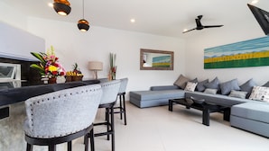 A modern living room with a grey sofa, a coffee table, and a bar area with two barstools. There is a painting on the wall and a ceiling fan. - Luxurious 4BR Villa With Pool, Bali Villa 2206 (Seminyak)