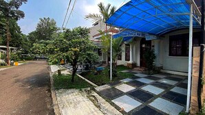 A residential area with a house featuring a blue awning, a paved driveway, and a small garden with palm trees. - Hotel O Pahlawan Udyana Syariah (Sentul City)