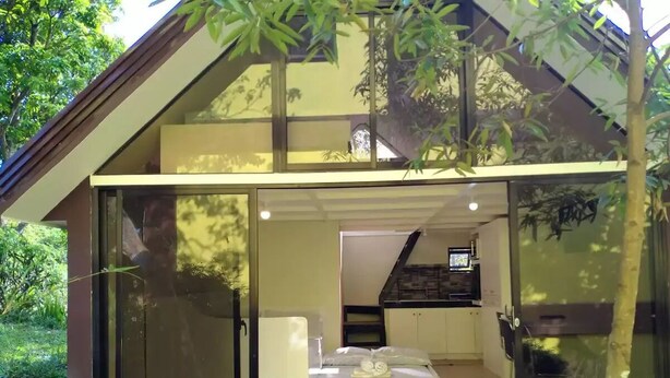 Una casa moderna con ingresso tramite porta scorrevole in vetro, una piccola camera da letto con un letto e un comodino, e un'area cucina visibile attraverso la porta.