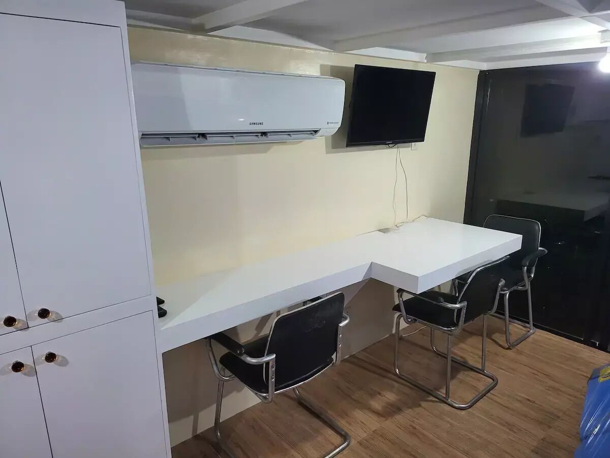 A compact room with a white desk, two black chairs, a mounted TV, and a white wardrobe.