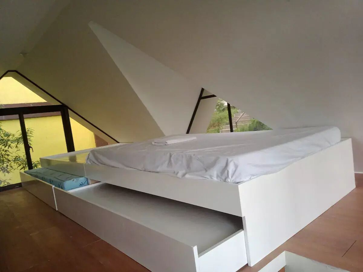 A modern loft-style bedroom with a built-in bed, a bench at the foot of the bed, and a large window providing natural light.