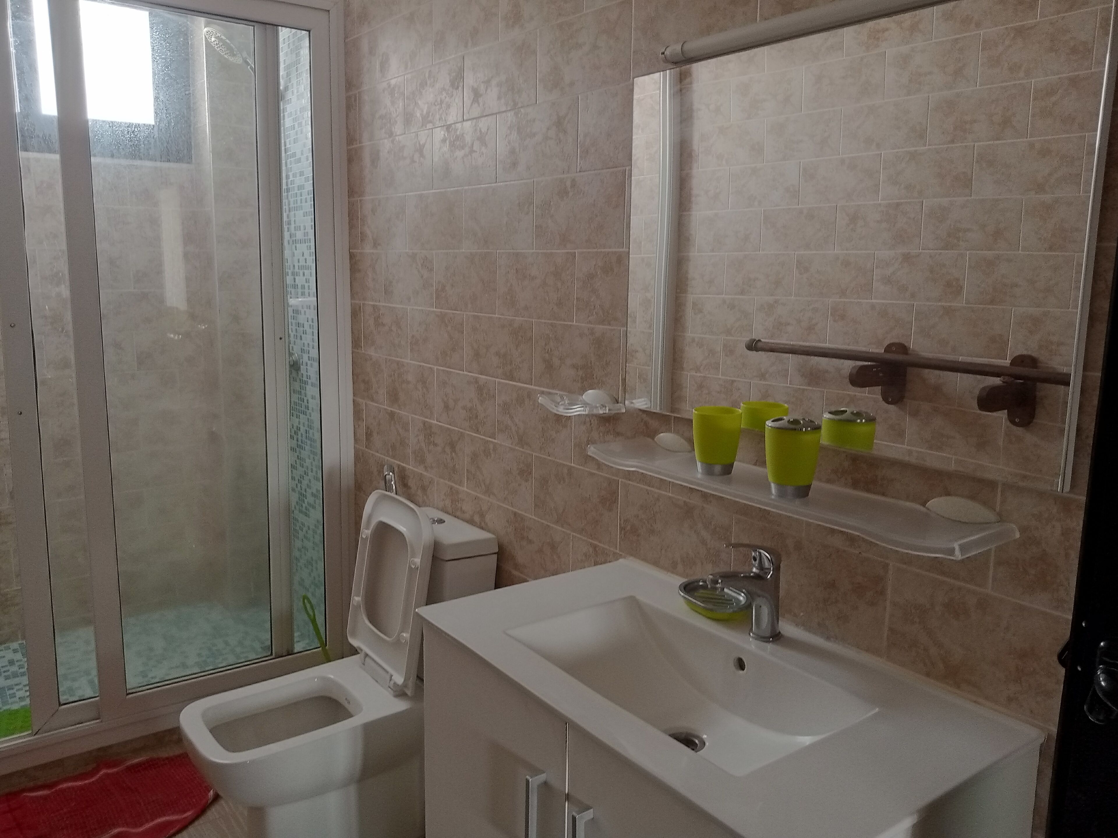 A bathroom with a toilet, sink, and shower. There are green cups on a shelf above the sink.