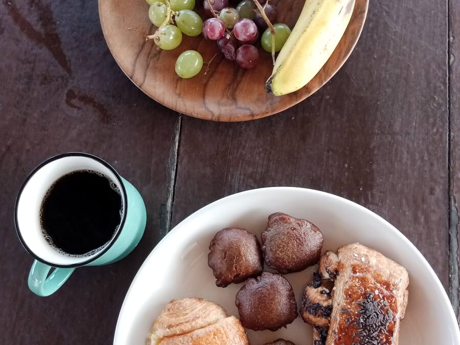 A wooden table with a bowl of mixed grapes and a banana, a mug of black coffee, and a plate with pastries and a grilled item.