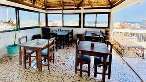 A restaurant with a thatched roof and tiled floor, featuring outdoor seating and large windows overlooking the city. - Residence Hotel Kakatar (Dakar)
