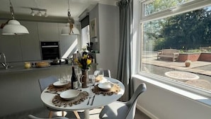 A dining area with a round table set for four, surrounded by grey chairs. A kitchen is visible in the background with modern appliances and a window overlooking a garden. - Sunny 2-Bedroom flat in Hoddesdon (Hoddesdon)