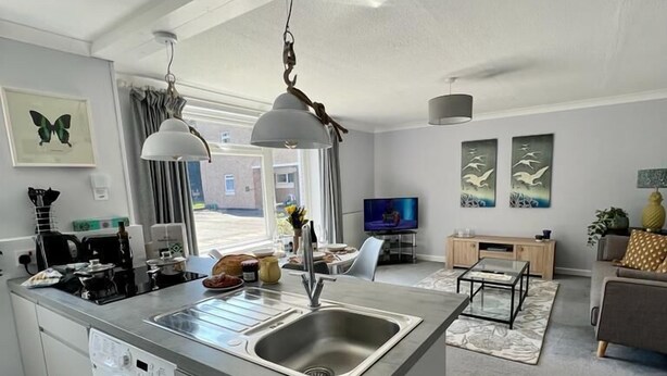 A modern kitchen with a built-in washing machine, a dining area with a TV, and a living room with a sofa and decorative items. - Sunny 2-Bedroom flat in Hoddesdon (Hoddesdon)