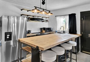 Une cuisine moderne dotée d’un réfrigérateur en acier inoxydable, d’un îlot central en bois avec des tabourets, d’un four à micro-ondes et d’un luminaire suspendu.
