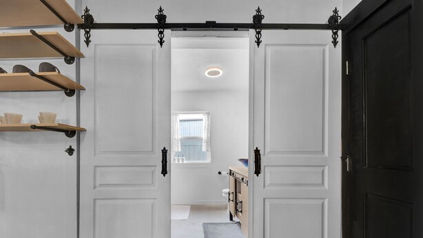 Un pasillo con una puerta negra a la derecha, una puerta blanca a la izquierda y una ventana que da a vistas exteriores.
