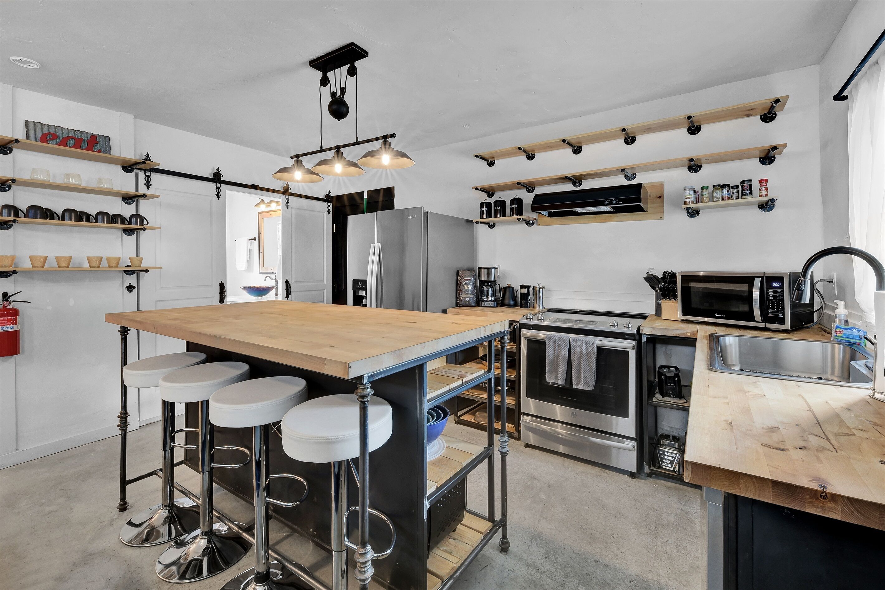 Una cocina moderna con una isla central, taburetes de bar, un refrigerador, un microondas y varios estantes con utensilios de cocina.