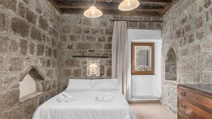 Een slaapkamer met een stenen muur, een bed met witte lakens, een houten dressoir en een klein raam met uitzicht.