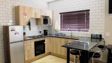 Cocina compacta con refrigerador, estufa, microondas y fregadero. Hay una ventana con persianas y un pequeño comedor con mesa y sillas.