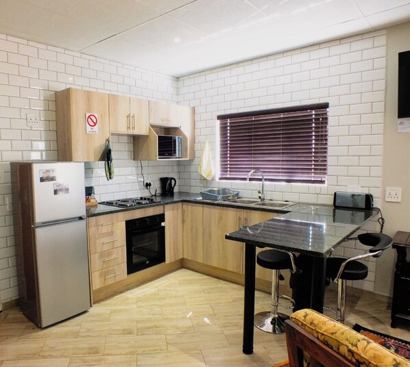A compact kitchen with a refrigerator, stove, microwave, and sink. There is a window with blinds and a small dining area with a table and chairs. - White Silo Accommodation (Thabazimbi)