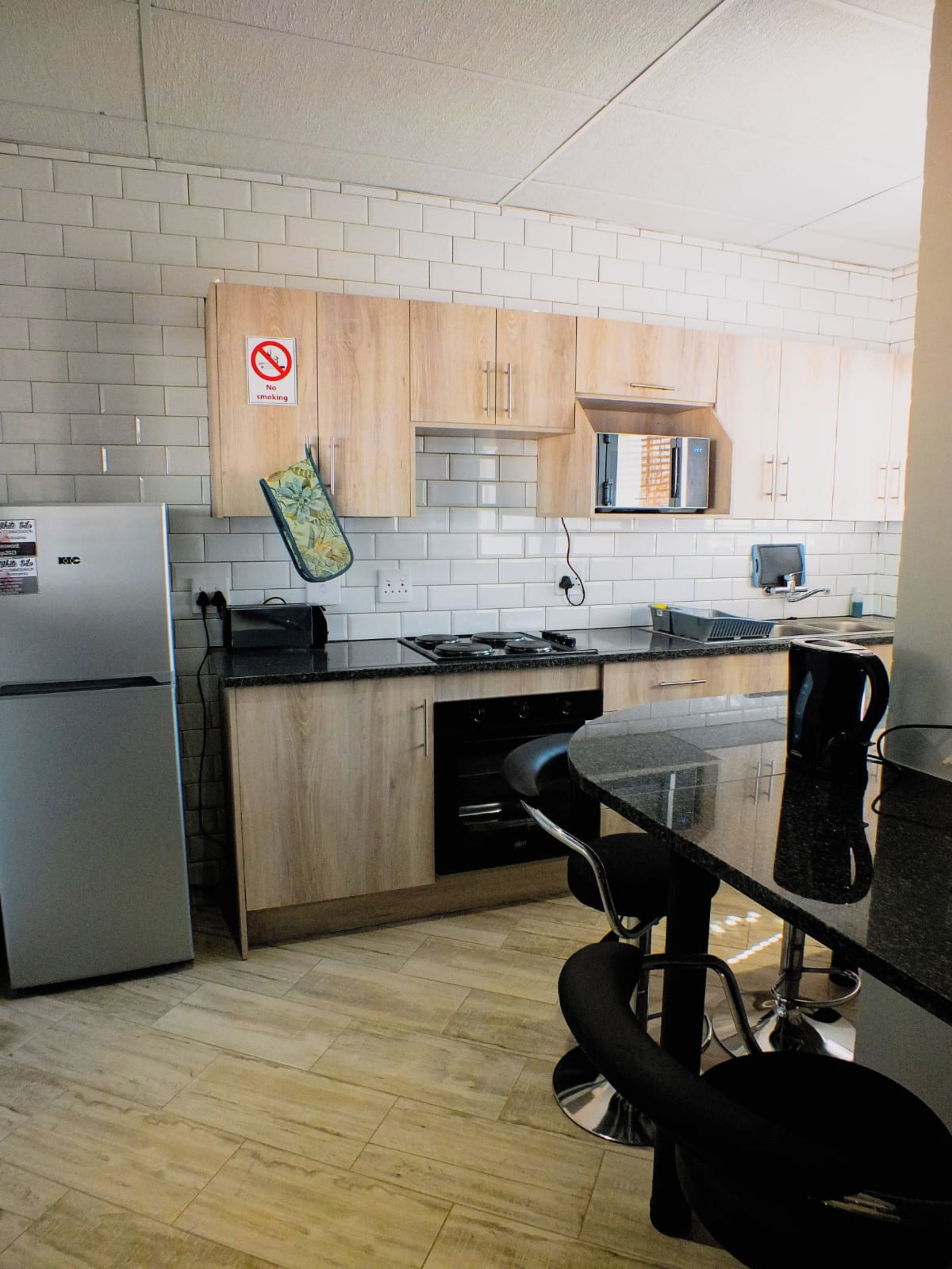 Una cocina con paredes de azulejos blancos, armarios de madera, un refrigerador de acero inoxidable, un microondas y una cafetera.