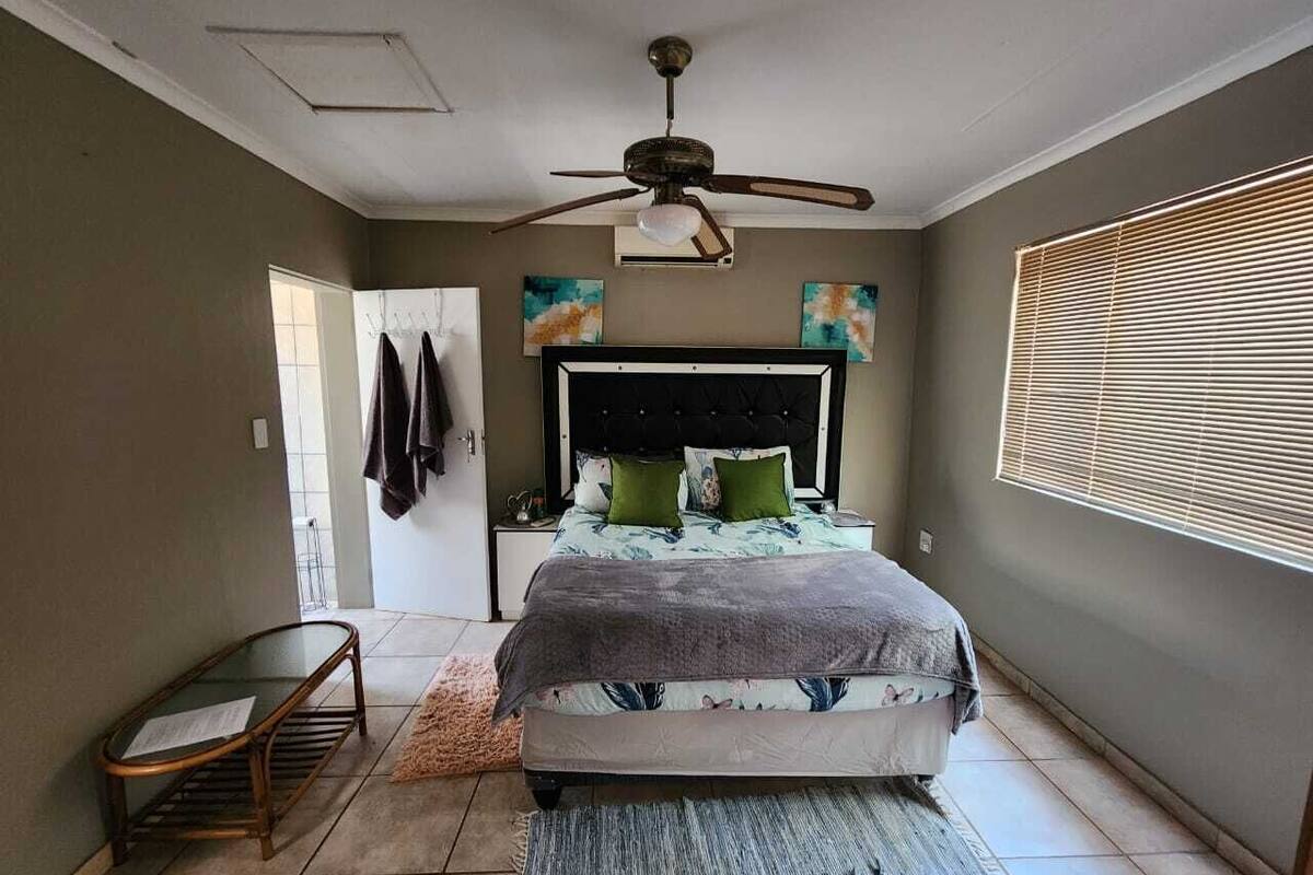Un dormitorio con cama, ventilador de techo, mesita de noche, una silla y ventana con persianas.