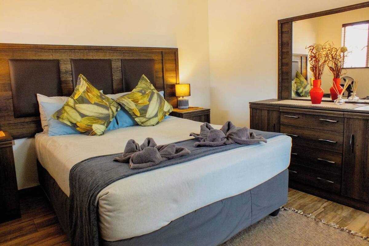 Una cama bien hecha con almohadas, una lámpara de noche, un aparador con cajones y un espejo.