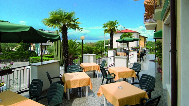 An outdoor patio with tables and chairs, palm trees, and a view of the sea. - hotel CASABLANCA (Lazise)