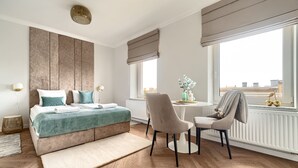 A modern hotel room with a large bed, a sitting area with a chair and table, a view of the city through large windows, and a radiator under the windows. - MARGI Daphne Apartment (Szczecin)