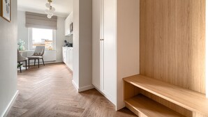 A hallway with a wooden floor, white walls, and a wooden bench. There is a window with blinds and a door with a handle. - MARGI Daphne Apartment (Szczecin)