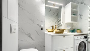 A modern bathroom with a white toilet, marble walls, a gold sink, and a built-in washing machine. - MARGI Daphne Apartment (Szczecin)