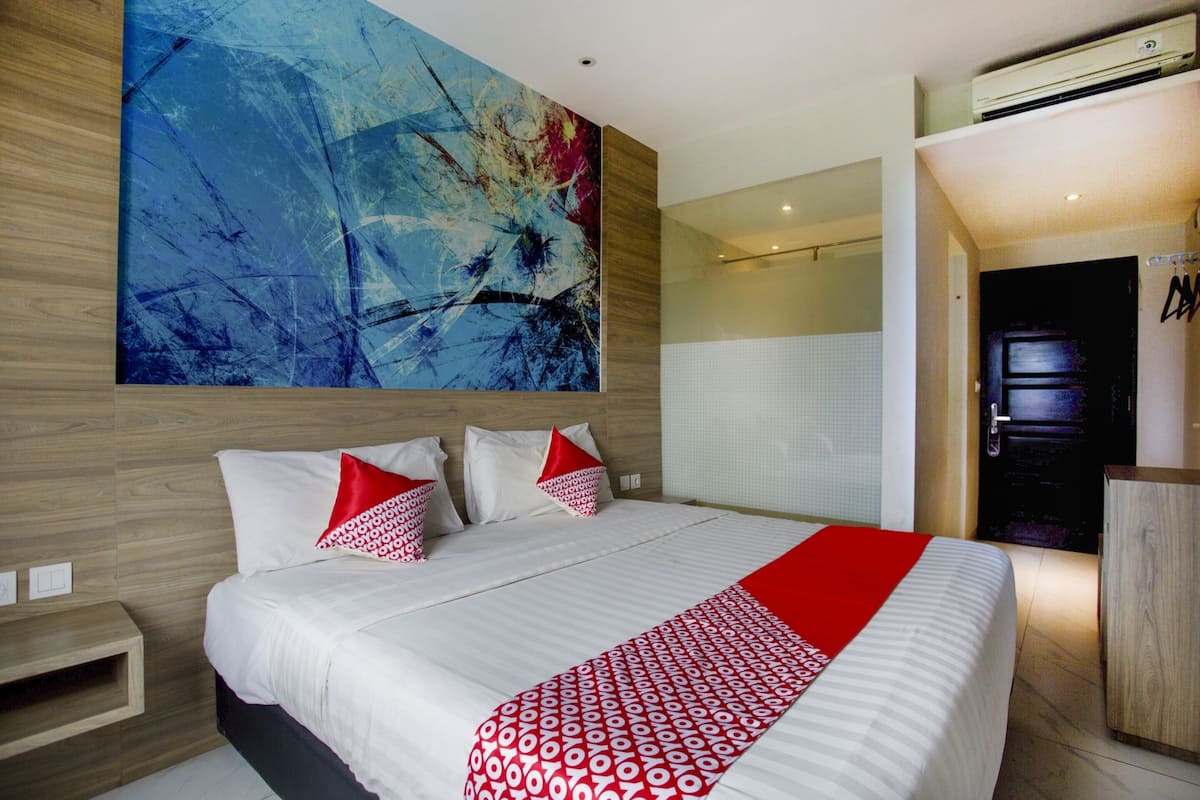 A modern hotel room with a large abstract painting on the wall, a neatly made bed with red and white pillows, and a red and white patterned throw.