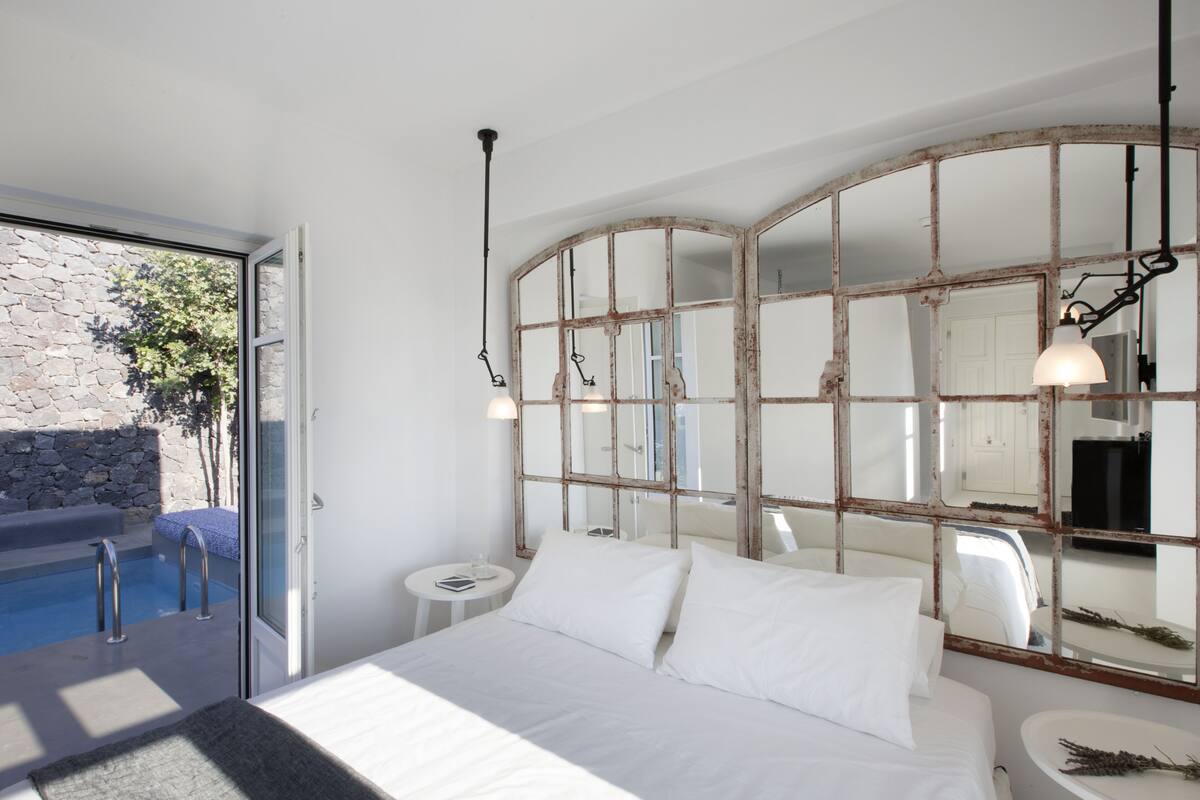 Quarto com uma cama grande, uma mesa pequena redonda, mesa de cabeceira, espelho grande e vista para uma piscina.