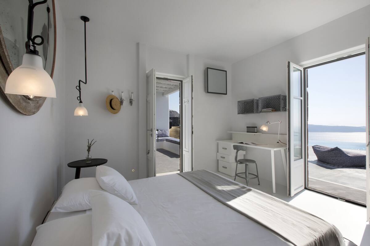 Quarto moderno com uma cama grande, uma escrivaninha e vista para o mar por uma porta aberta.