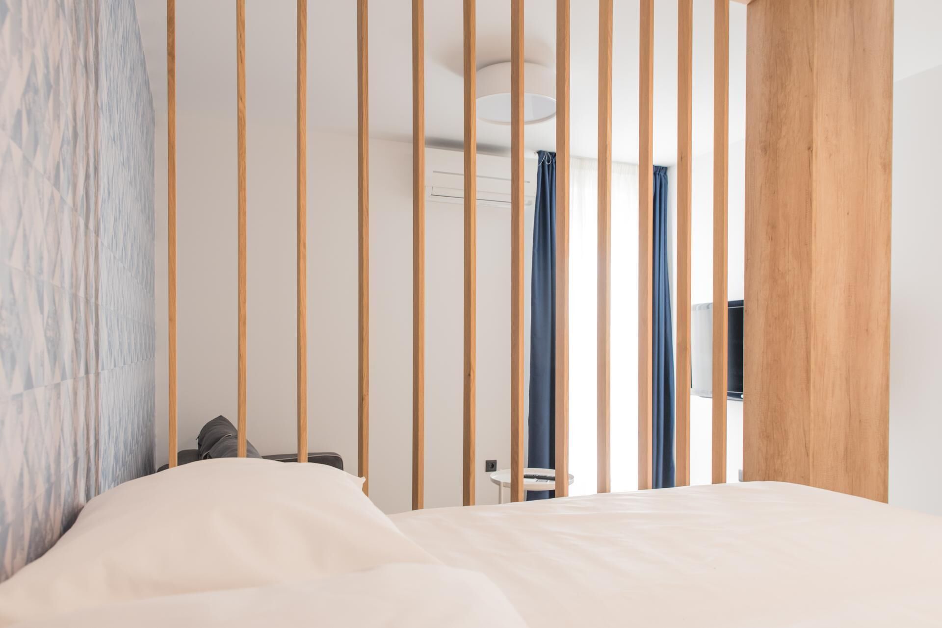 Habitación de hotel con cama, una mesita y una silla. Se observa un separador de madera con listones verticales, una pared con textura y una lámpara en el techo.