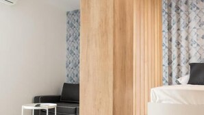 A modern hotel room with a wooden accent wall, a bed with a white mattress, a small white side table, and a patterned curtain. - Deluxe, Charming and Cozy Bcadria Apartment A2 (Split)