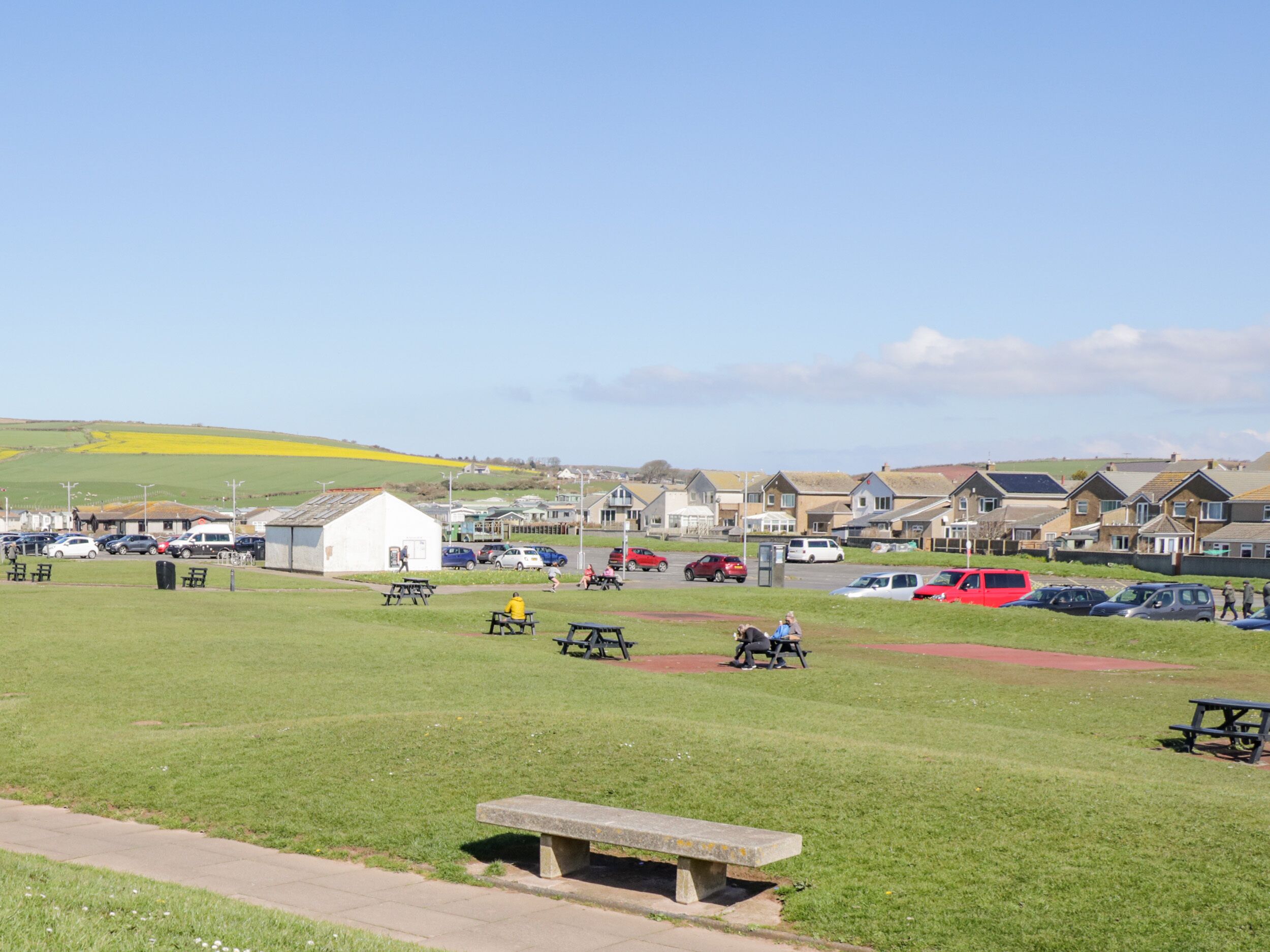 A grassy area with picnic tables and benches, a small white building, and a row of houses in the background.
