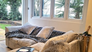 A cozy living room with a sofa, a fur throw, a book, and a champagne bottle in a bucket. - 1 Bed Cottage-museum View-garden-free Parking (Weybridge)