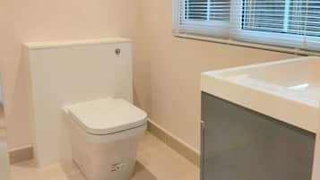 A compact bathroom with a toilet, a washing machine, and a window with blinds.