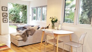 A cozy living room with a sofa, a small round table, and chairs. There are framed pictures on the wall and a window with a view of greenery outside. - 1 Bed Cottage-museum View-garden-free Parking (Weybridge)