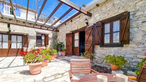 Un bâtiment en pierre avec des volets en bois, une table en bois sur laquelle sont posées deux tasses blanches, et des plantes en pot.