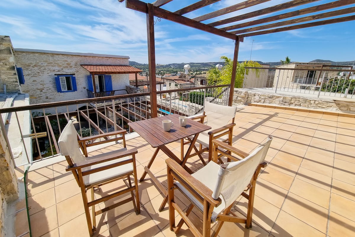 Eine Holz-Pergola mit Tisch und Stühlen auf einer gefliesten Terrasse mit Blick auf ein Wohngebiet mit Steinhäusern und blauem Himmel.