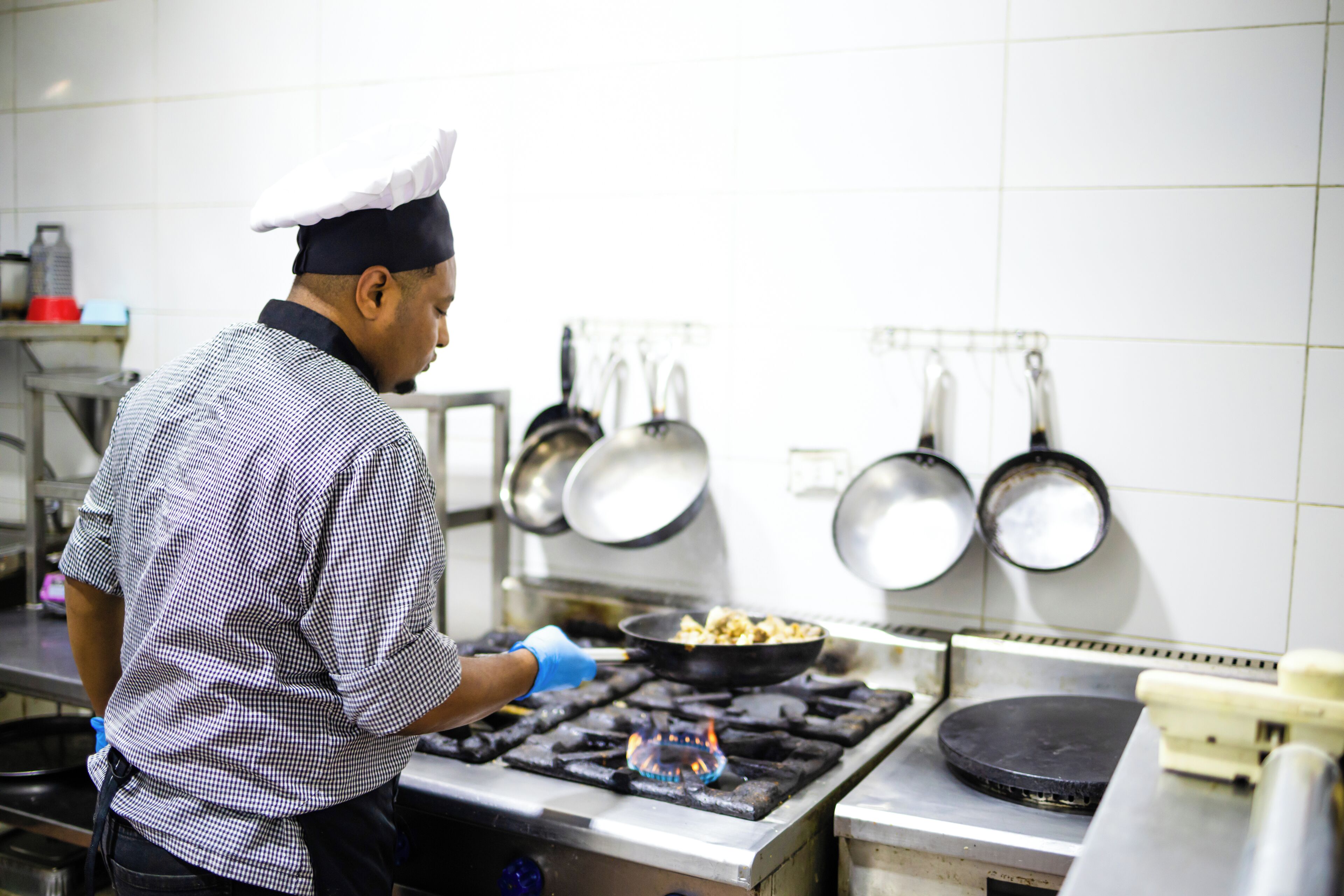 Een kok kookt in een keuken met roestvrijstalen apparatuur en hangende pannen.